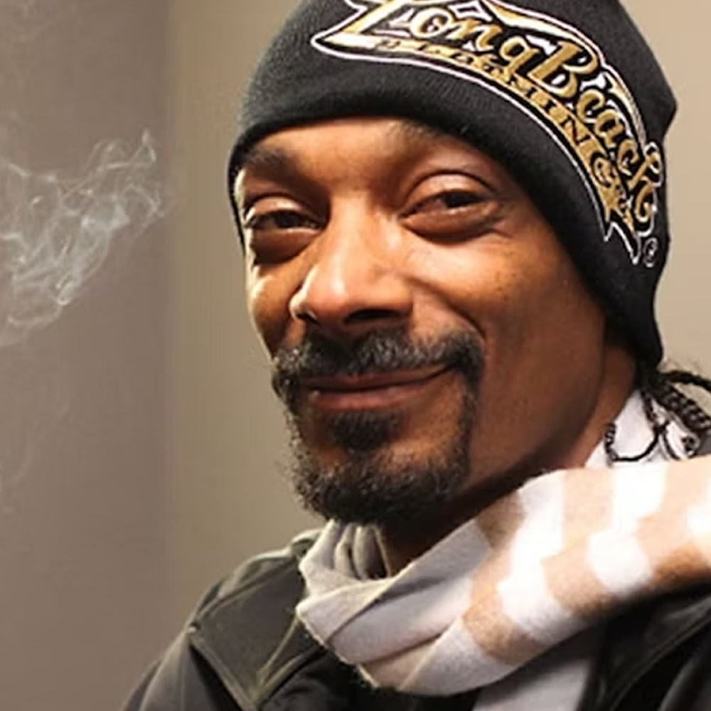 Snoop Dogg, head and shoulders, smiling slightly at the camera. He wears a black knitted beanie reading "Long Beach" in gold, has a goatee, and a patterned scarf at his neck. A wisp of smoke drifts in the left of the frame against a plain grey wall.