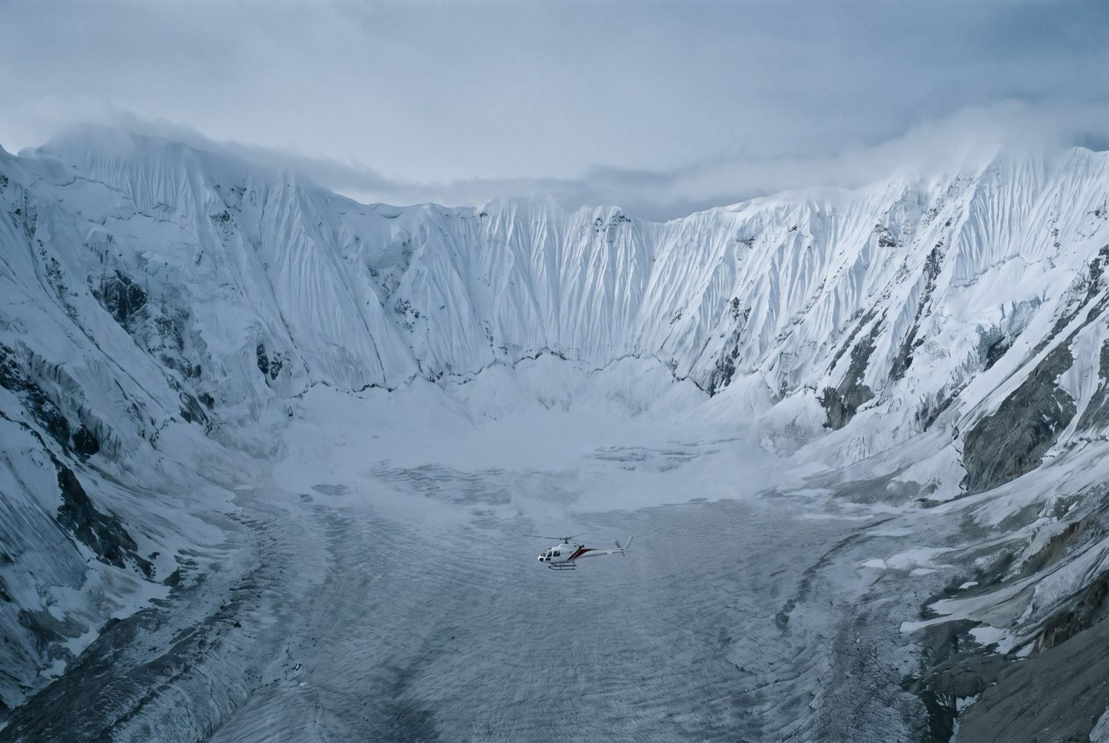 A minute white helicopter crossing the floor of a vast glacial basin entirely ringed by sheer fluted snow walls.