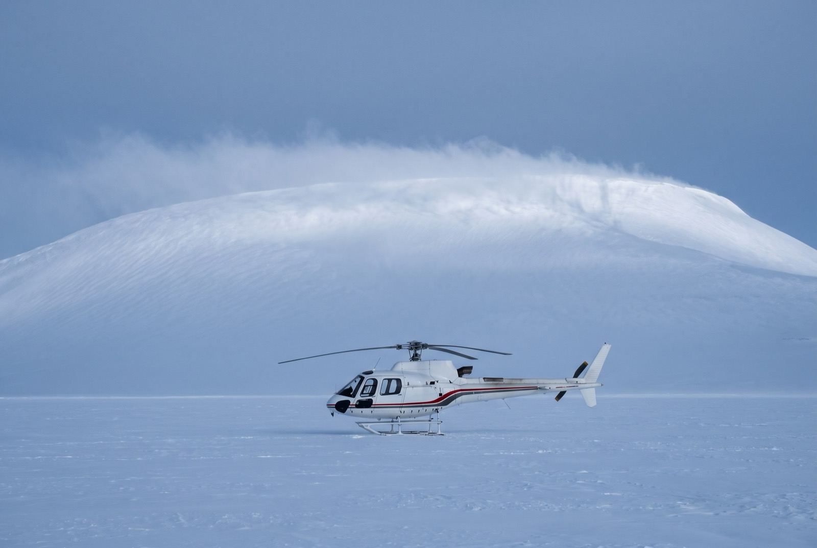A white helicopter alone on a vast flat snow plateau beneath a broad whaleback snow dome trailing wind-blown snow.