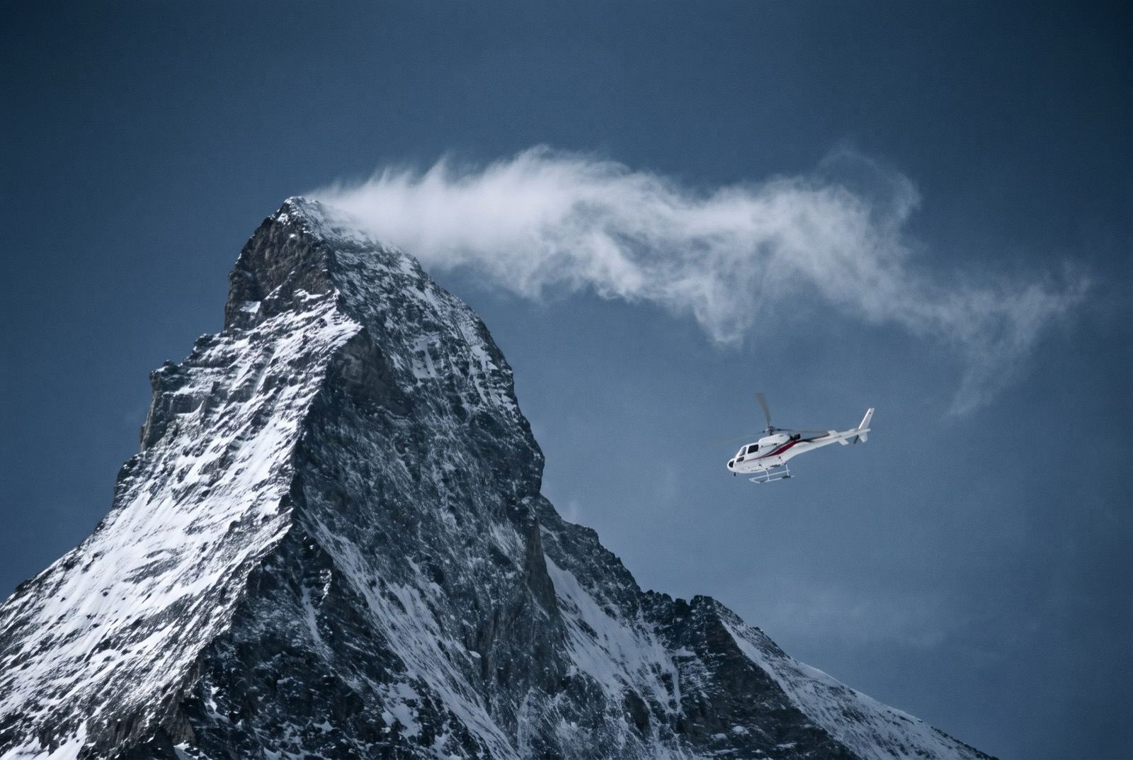 A small white helicopter at extreme altitude near a dark summit pyramid trailing a long horizontal banner of spindrift.