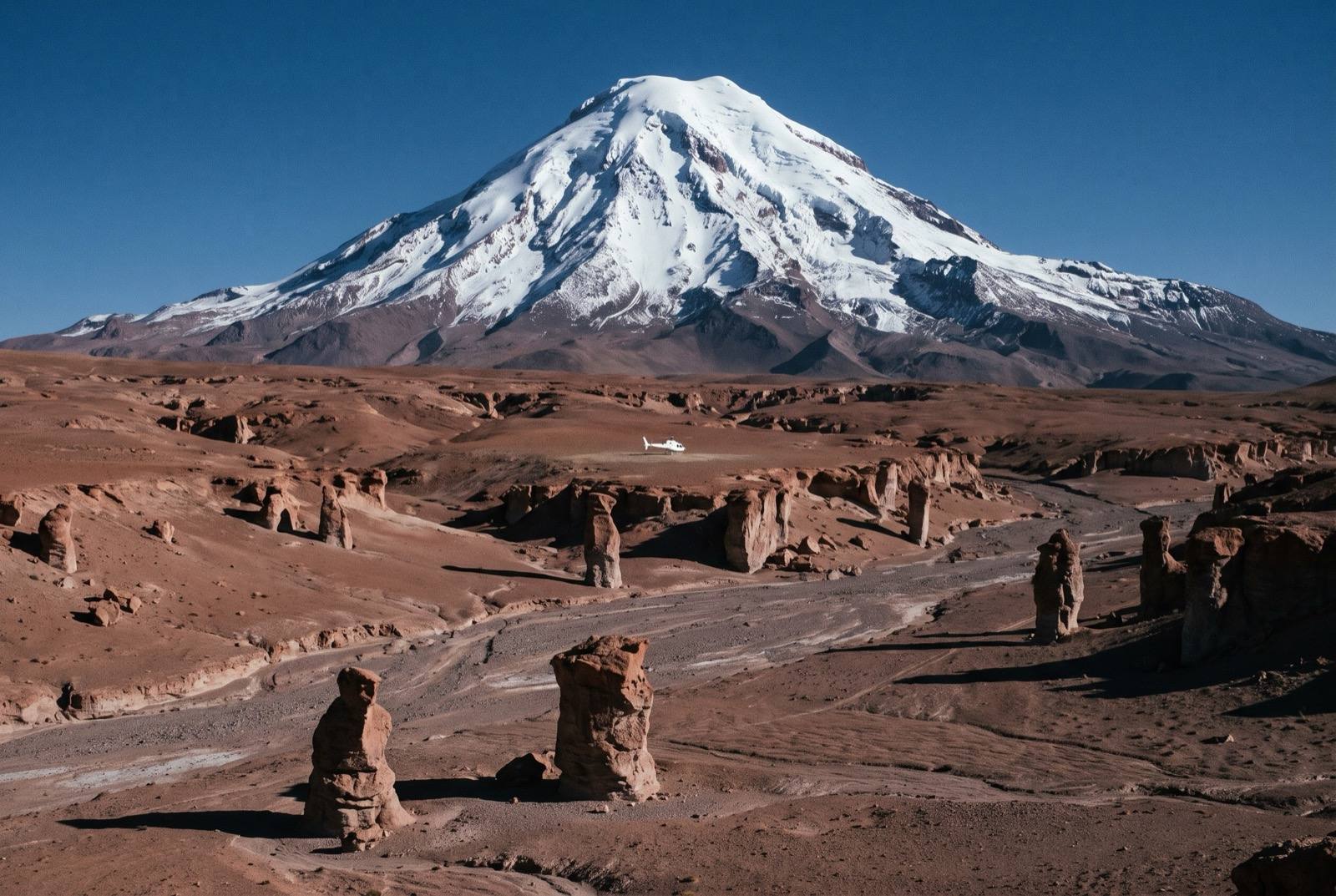 A small white helicopter standing far off on a bare brown high-desert plateau below a single white glaciated peak.