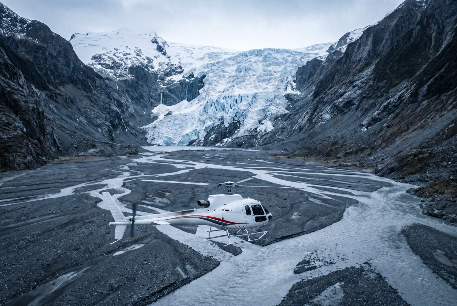 A white helicopter standing on braided grey river flats below a hanging icefall in a broad glaciated valley.