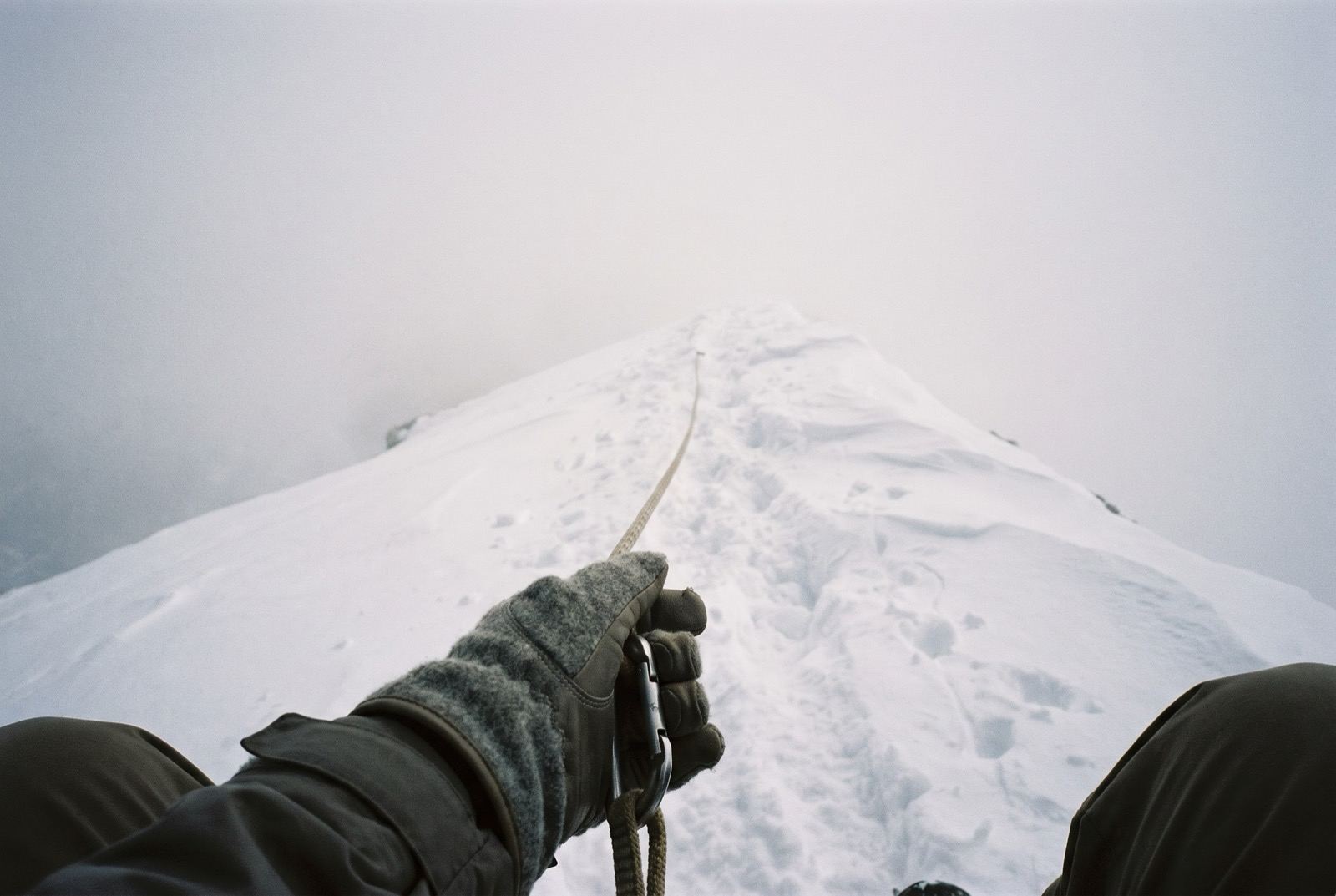 A gloved hand clipped to a fixed line running along a corniced ridge that vanishes into flat grey cloud.