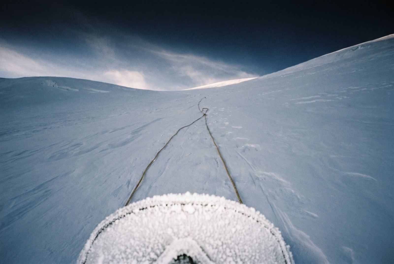 The frost-encrusted top edge of an oxygen mask at the bottom of the frame, two rope strands running up a pale snow slope beyond.