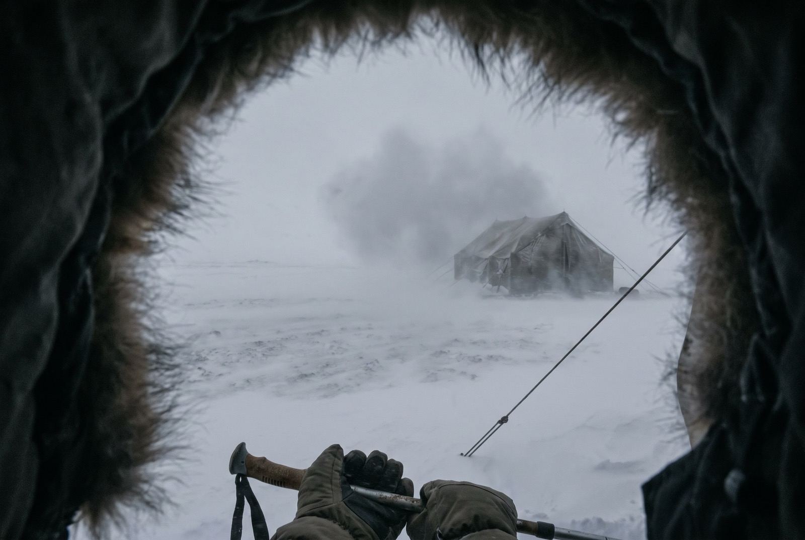 The fur-trimmed edge of a hood framing the view, gloved hands on trekking poles, a tent and a guy-line dim in grey cloud.