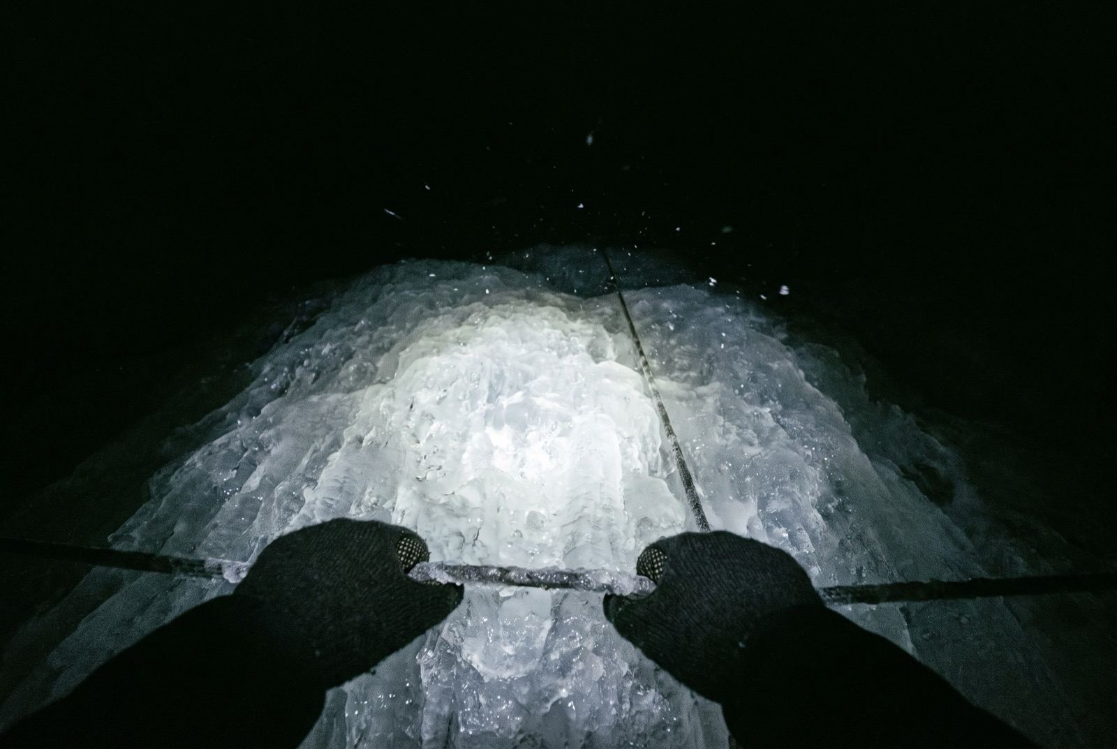 Two gloved hands on a fixed rope at the bottom of the frame, a head-torch cone lighting steep ice a metre ahead, blackness beyond.