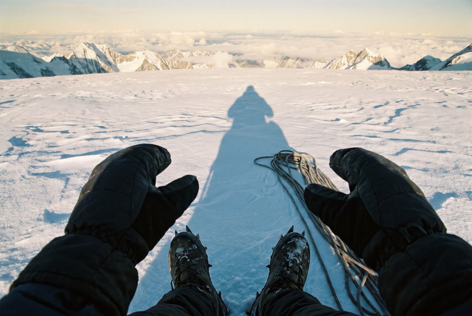 A long shadow thrown across untouched summit snow with boots and a coiled rope at the bottom of the frame, distant peaks on the horizon.