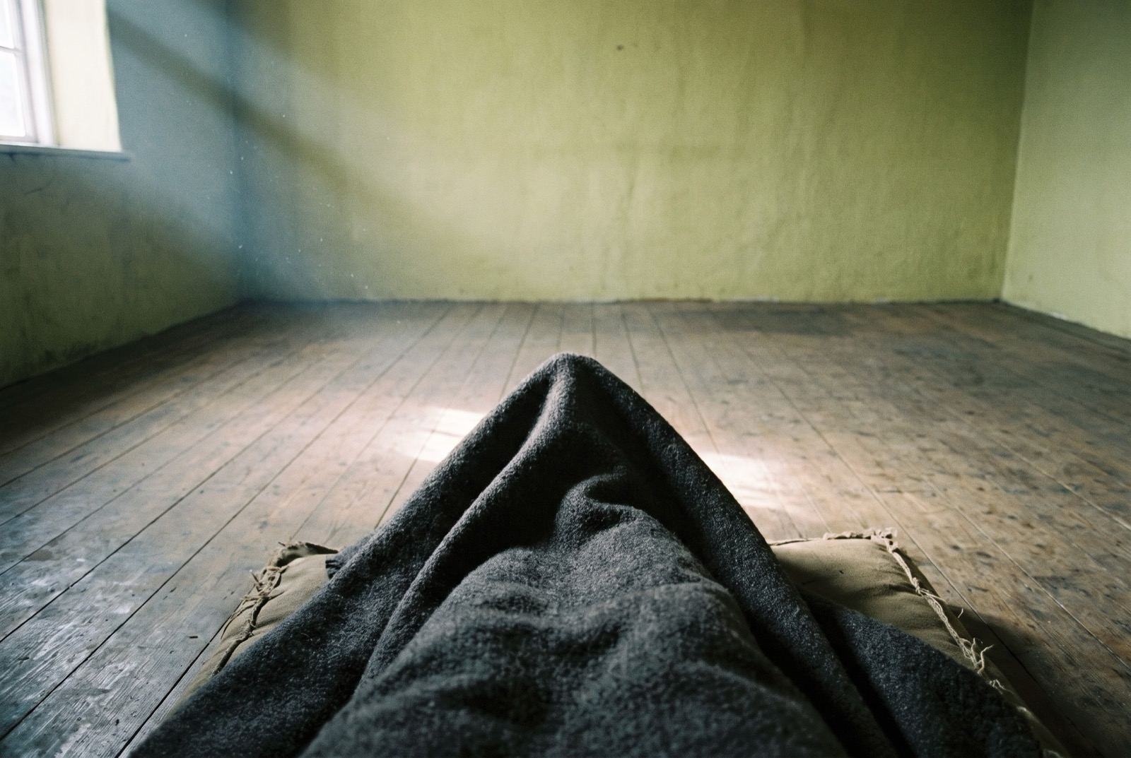 Looking down at crossed legs under a plain dark blanket on bare floorboards, dust turning in a shaft of cold light.