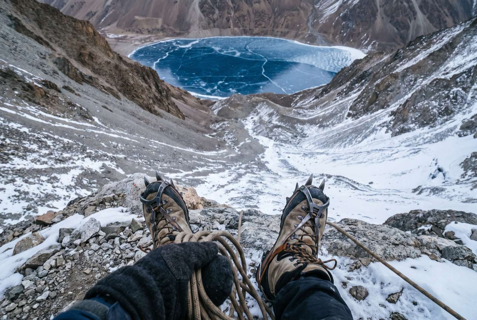 Looking down past crampons on broken moraine to a frozen blue lake far below in a bowl of rock.