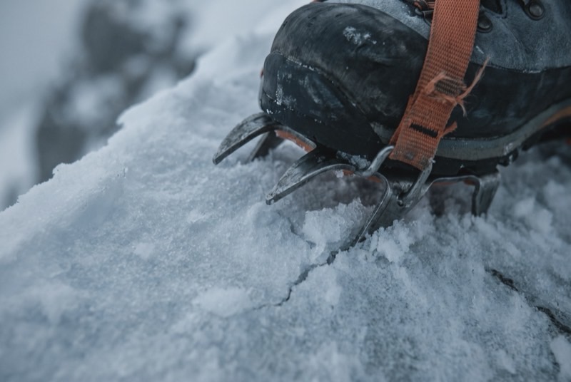 Crampon front points biting into wind-hardened snow on a ridge crest, an orange gaiter strap just in frame