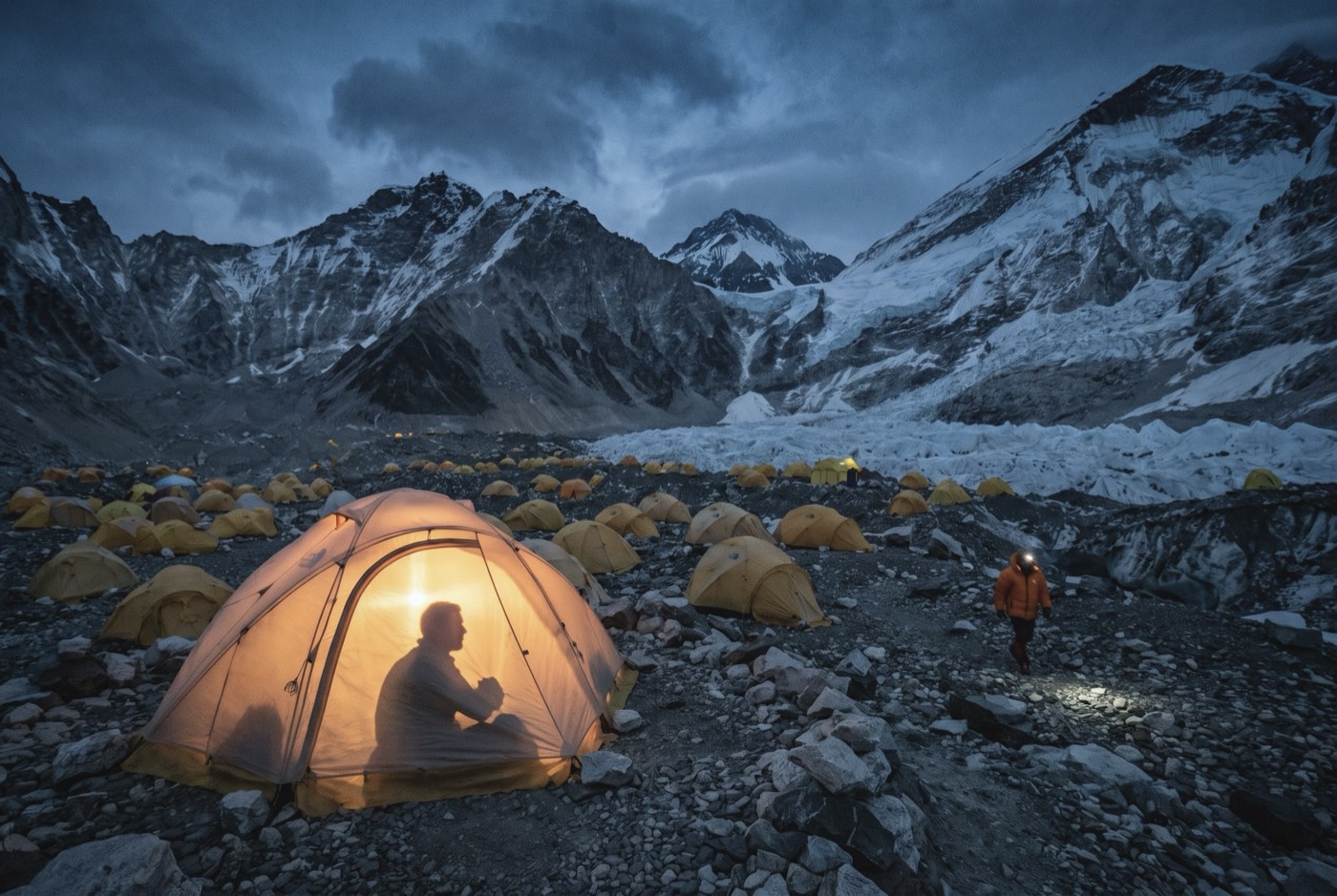 Everest Base Camp at blue hour: hundreds of yellow tents across the Khumbu glacier, one lit from inside with a figure sitting up in it, and a climber in an orange jacket walking between them by head torch.