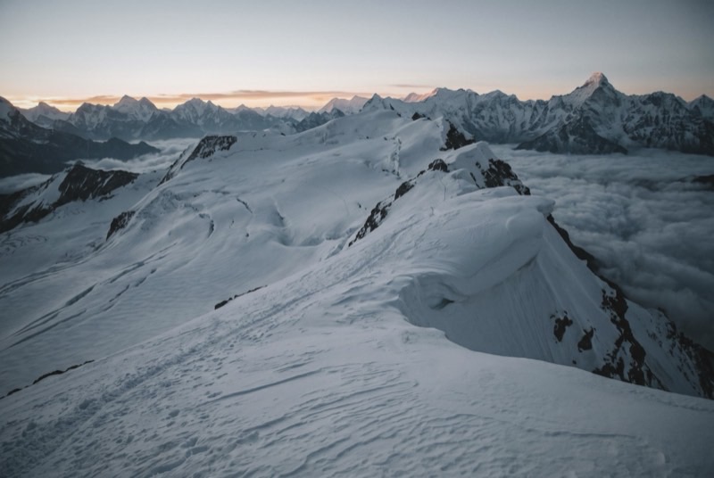 An empty glaciated snow ridge with a curling cornice, above a sea of valley cloud at dawn.