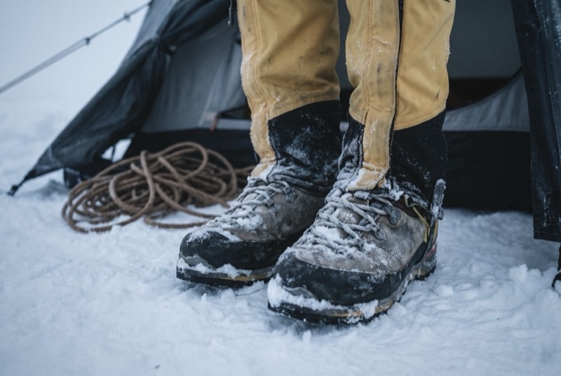 Frost-rimed mountaineering boots with yellow gaiters standing in packed snow outside a tent door.