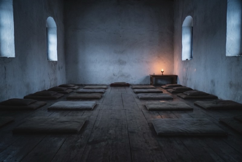 Rows of empty cushions on the worn wooden floor of a deserted monastery meditation hall at first light, a single butter lamp burning against the far wall.