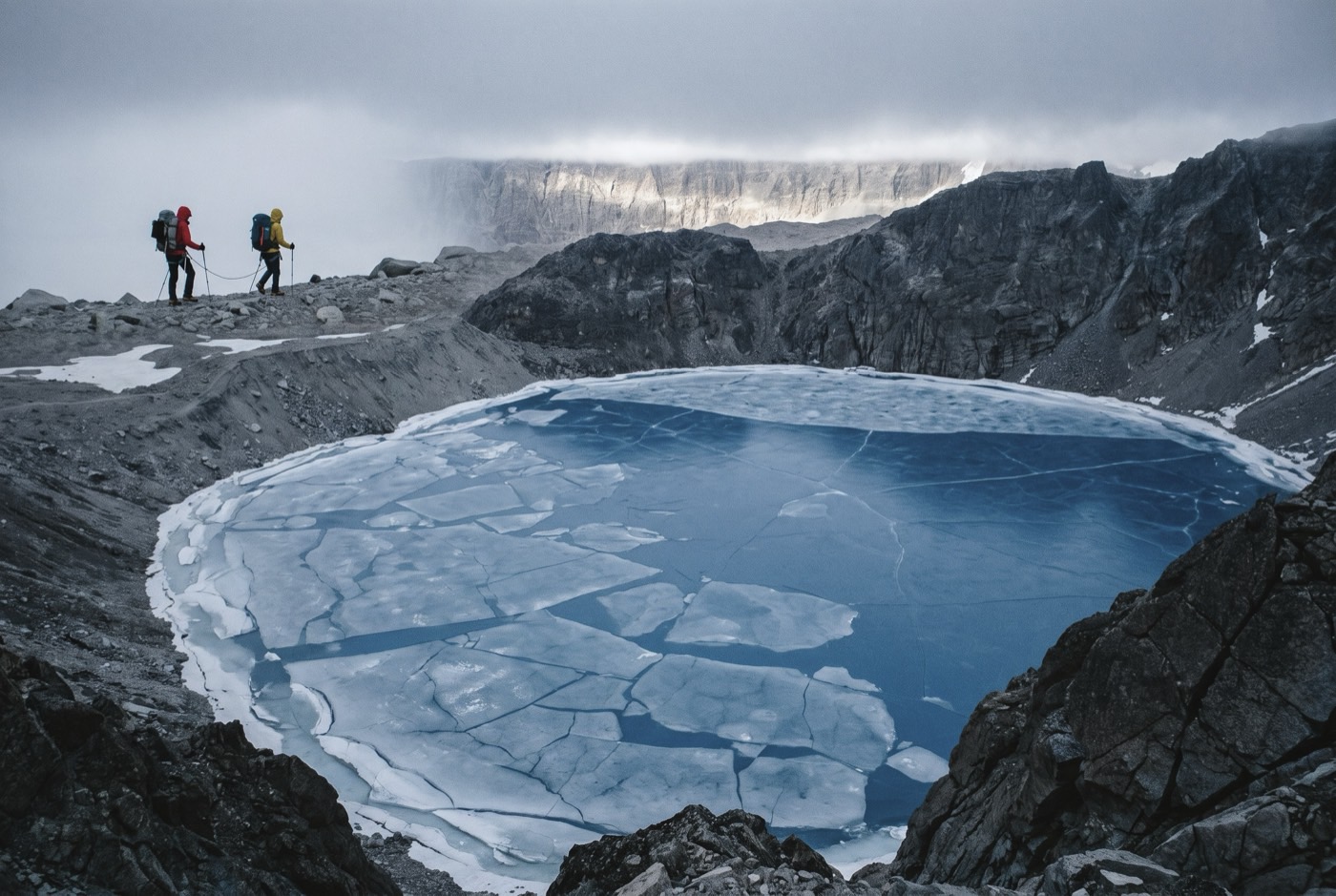 A rope team of two crossing the moraine high above the frozen cobalt ice of Tilicho Lake, tiny against the rock bowl